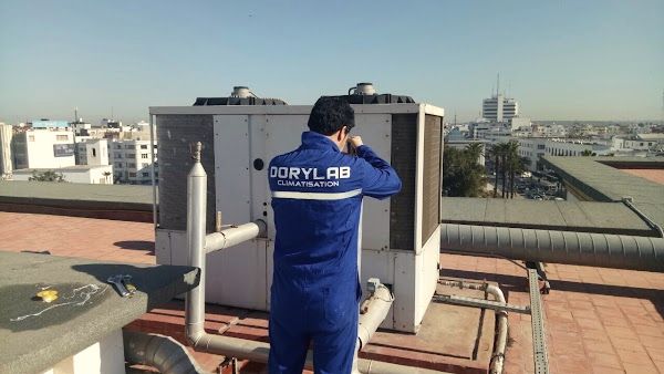 Technicien en combinaison bleue Dorylab, de dos, travaillant sur une unité de climatisation en toiture, la ville en arrière-plan