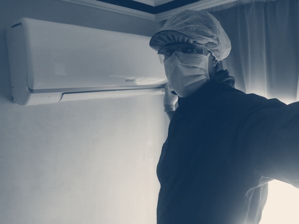 Technicien en charlotte, lunettes, masque et gants, à côté d’un climatiseur mural